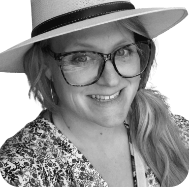 Une femme souriante portant des lunettes et un chapeau à larges bords dans un portrait en noir et blanc.