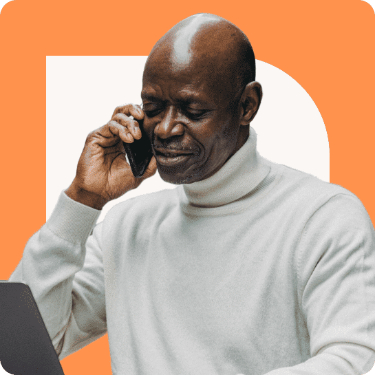 Un homme noir plus âgé vêtu d'un pull blanc parle sur un smartphone, avec un ordinateur portable ouvert devant lui, sur un fond orange.