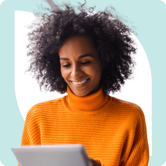 Une femme aux cheveux bouclés, vêtue d'un pull orange, souriant en regardant une tablette.